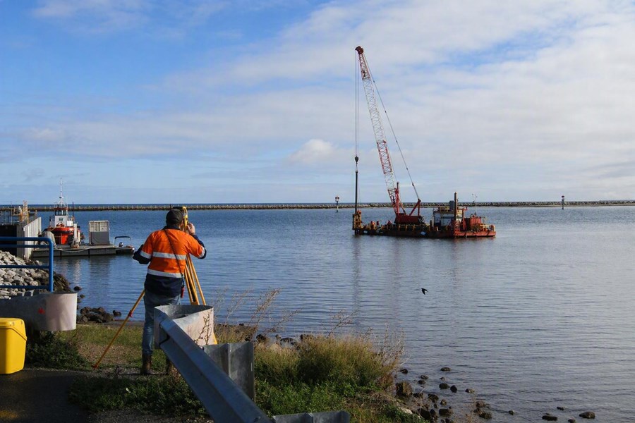 The image shows marine works being carried out at Outer Harbor, South Australia, near the entrance to the Port River and the Royal South Australian Yacht Squadron (RSAYS). In the foreground, a surveyor stands on the shoreline operating a surveying instrument mounted on a tripod. The surveyor is positioned beside a paved waterside path, with safety barriers, rocks and coastal vegetation along the shoreline. On the water, a floating work barge supports a large crane with a tall lattice boom extending high into the sky. The crane appears to be engaged in marine construction or maintenance work within the harbour. Several navigation markers are visible in the water behind the barge, helping guide vessel traffic through the harbour entrance. To the left of the image, a small service vessel is moored alongside a floating pontoon. In the background, a long breakwater stretches across the horizon, protecting the harbour from offshore swell. The water is calm, reflecting the overcast sky above. The photograph highlights surveying and construction activities associated with maintaining marine infrastructure at the entrance to the Port River.
