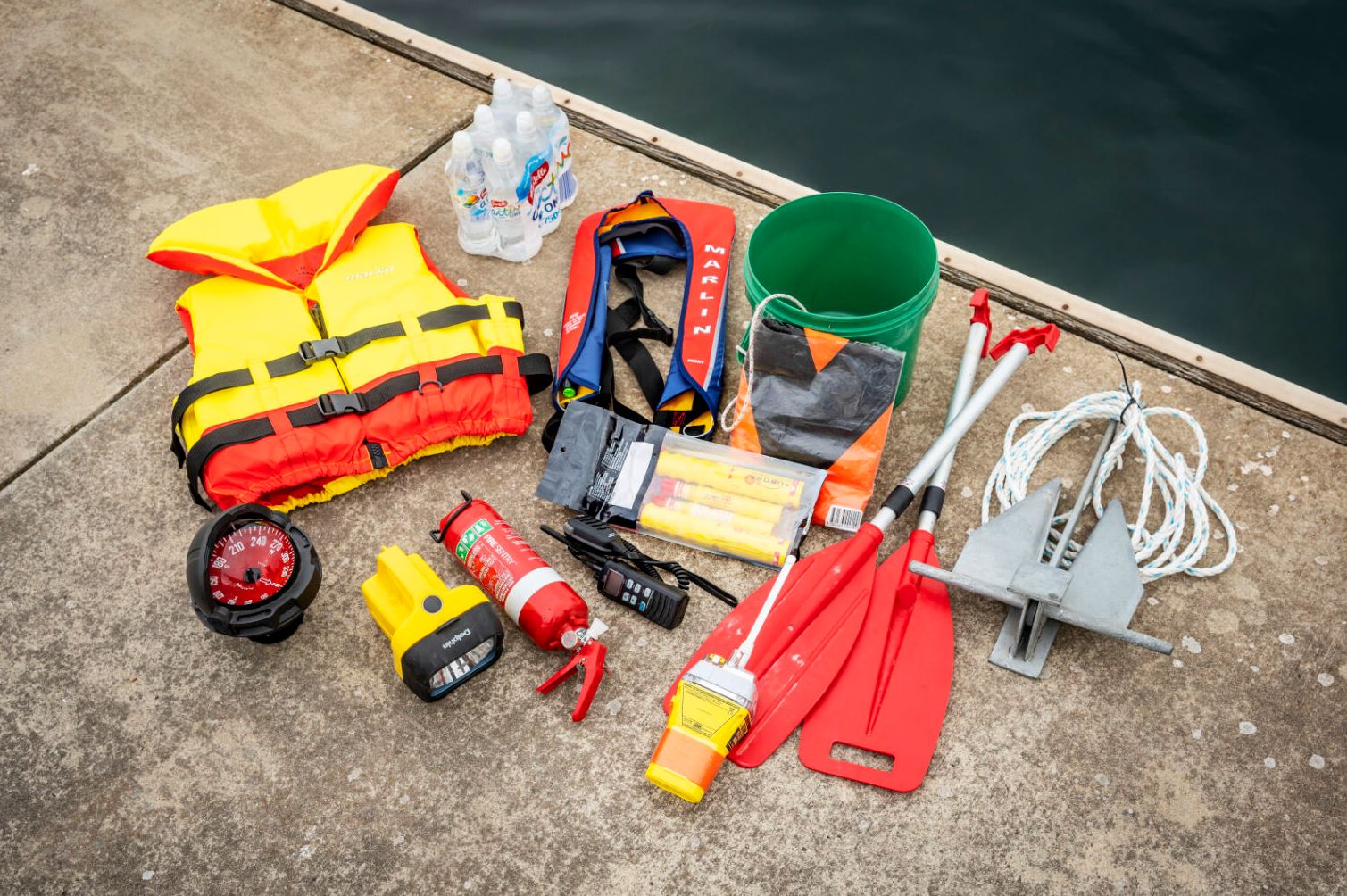 Various safety gear on the ground - lifejacket, compass, torch, fire extinguisher, flares, radio, bucket, V sheet, EPIRB, paddles, anchor 