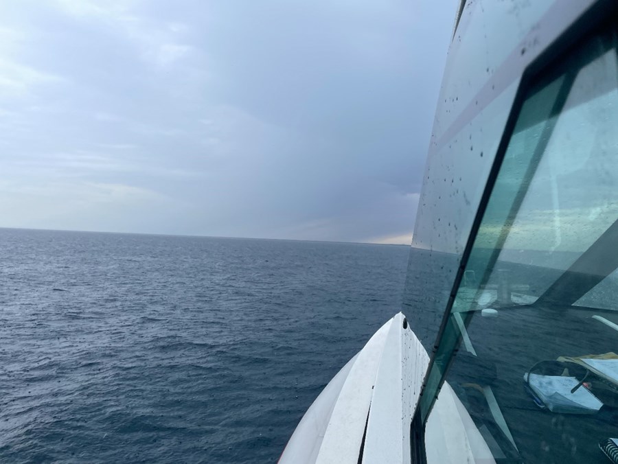 The image is taken from aboard Wardana, a Marine Safety SA vessel operating at sea. The photograph looks forward along the starboard side of the vessel, with part of the white wheelhouse and large cabin windows visible in the foreground. Water droplets can be seen on the windows, suggesting recent rain or rough weather conditions. The vessel is travelling across open water under a dramatic sky filled with dark storm clouds. The sea is a deep blue-grey, with small waves and ripples extending to the horizon. In the distance, a faint strip of land is visible beneath the cloud cover. The image highlights the challenging conditions that boaters may encounter while conducting activities on South Australian waters.