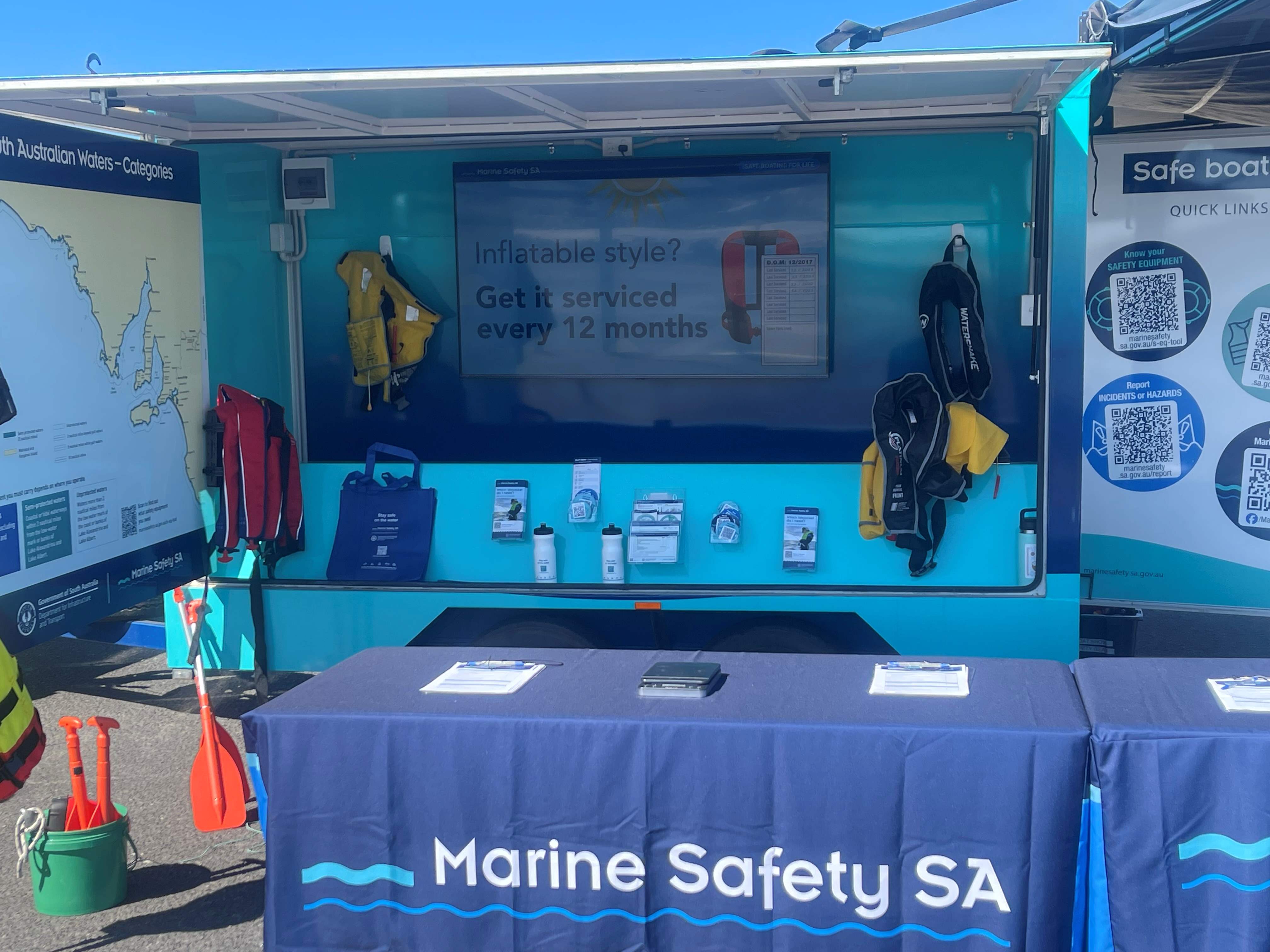 Pictured is our Marine Safety Stay Afloat Trailer where we conduct our Stay Afloat clinics for general public located in different areas across the state. Pictured is our trailer opened up with view of the tv hung from the back wall with key safety messages. Brochures are in the image alongside our Marine Safety drink bottles and lifejackets