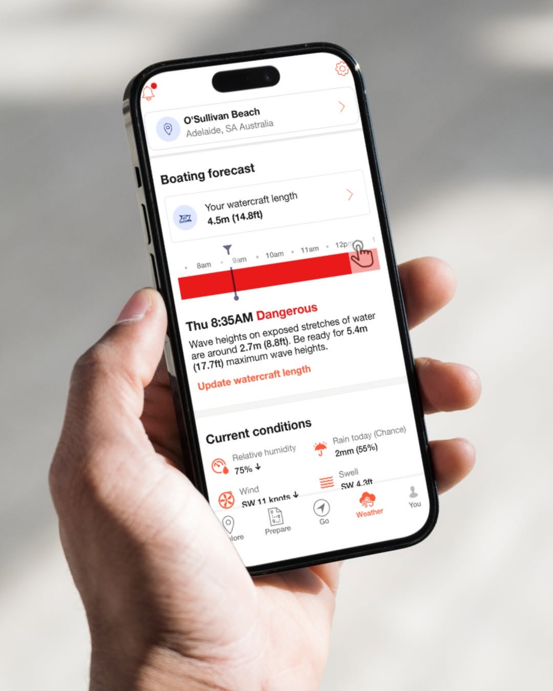 The image shows a smartphone displaying the Deckee boating safety app. The app is open to a location at O'Sullivans Beach, Adelaide, South Australia, with vessel details entered and boating conditions displayed for the day. A prominent warning on the screen indicates that conditions are considered dangerous at a specific time, alerting boaters that waves on exposed stretches of water may exceed the safe operating limits for the selected vessel. Additional information shown within the app includes wind speed and direction, swell conditions, and other marine weather data to help boaters assess conditions before heading out on the water. The smartphone is held in one hand against a blurred outdoor background, emphasising the convenience of accessing important safety information while on the go. The image highlights how the Deckee app can help recreational boaters make informed decisions and improve safety on the water.