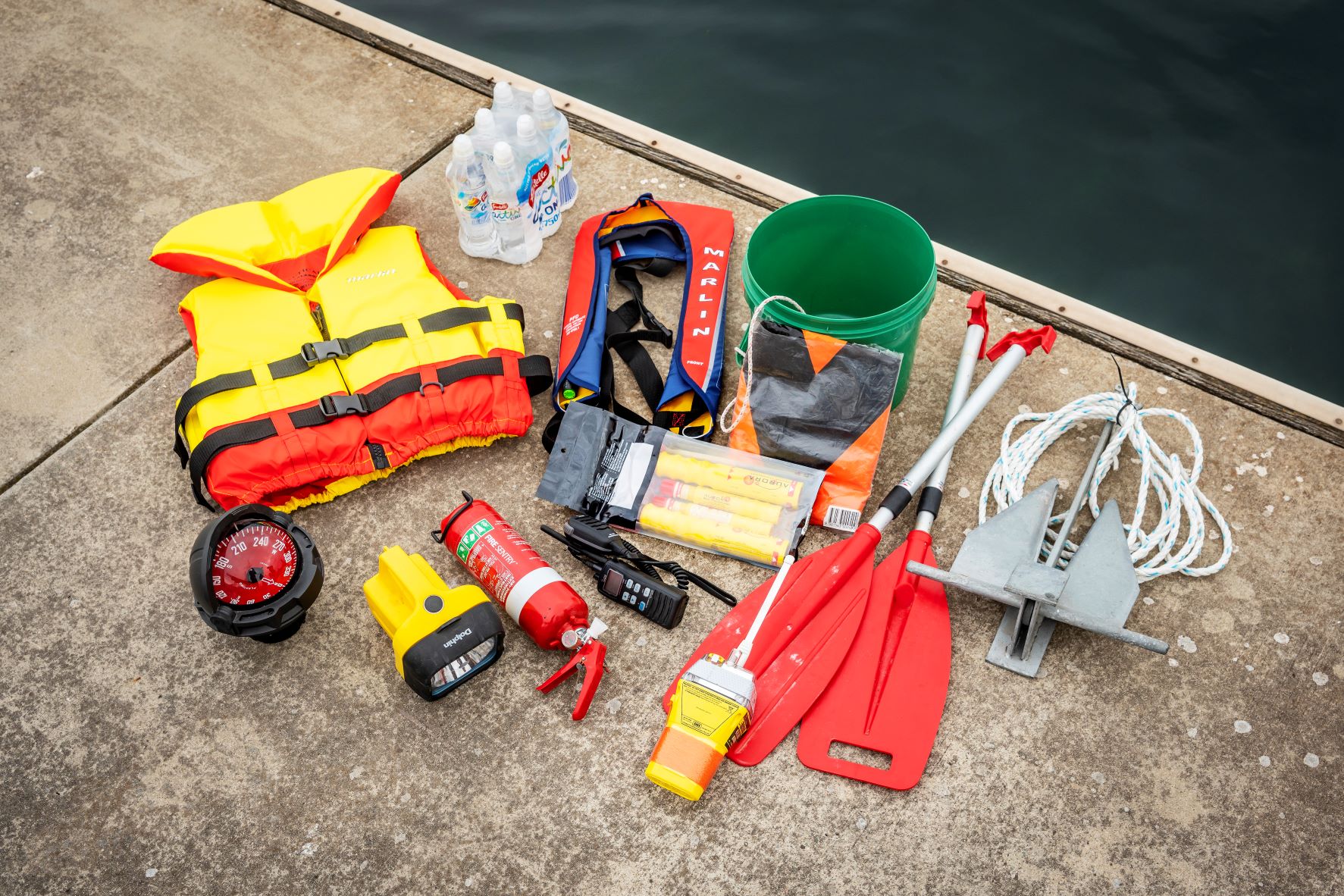 Range of safety equipment laid out on a wharf - lifejacket, compass, torch, fire extinguishers, EPIRB, paddles, flares, radio, anchor, bucket, V sheet, bottled water